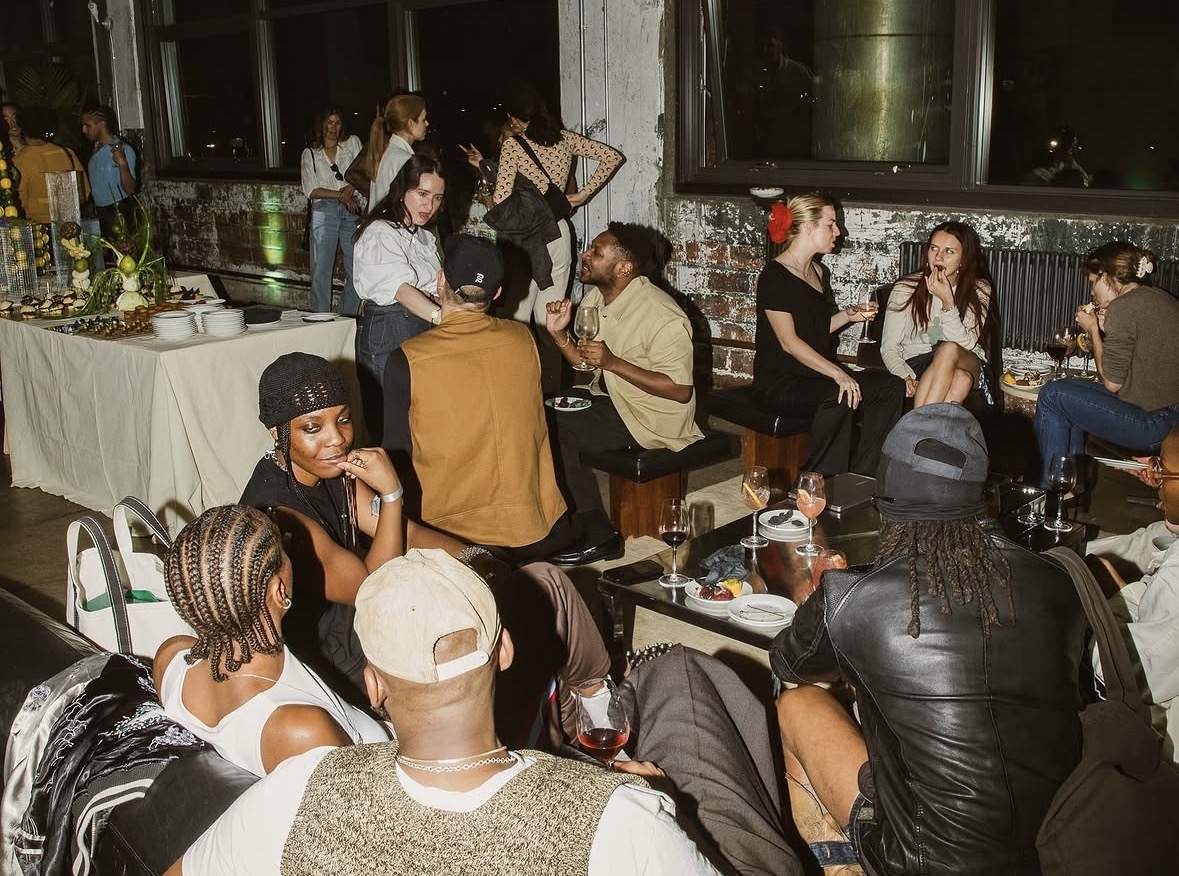 A crowded indoor party with guests holding wine glasses around low tables, seated and standing against exposed brick walls, with a buffet table of plates and food at the back left