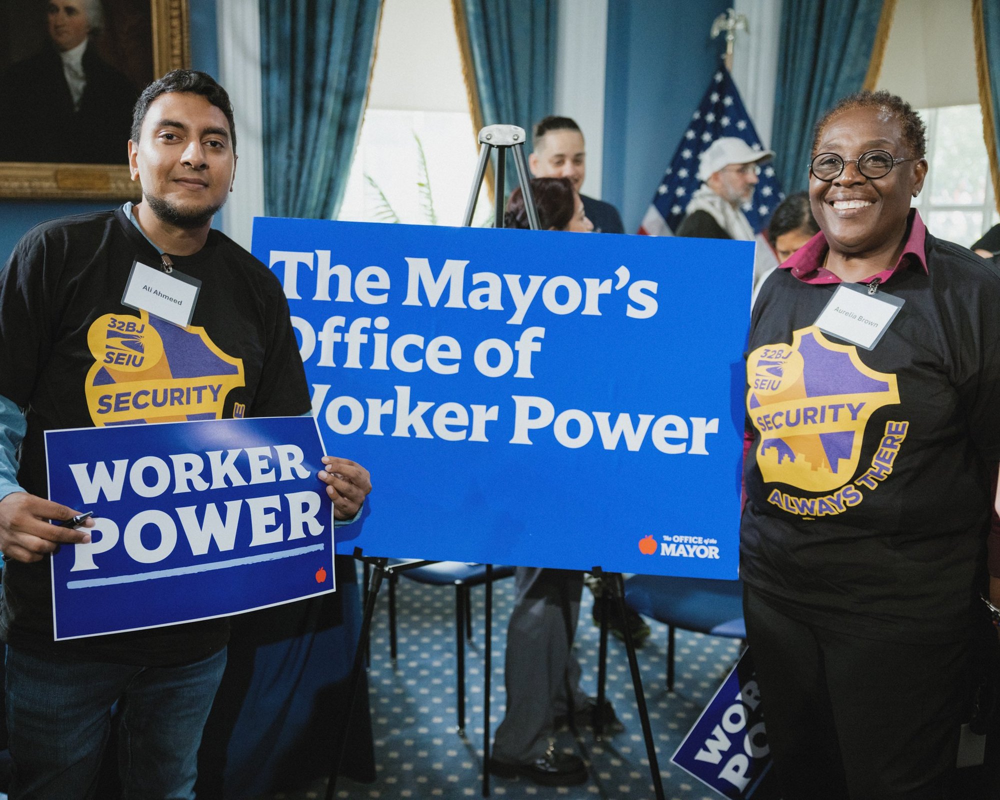 Two 32BJ SEIU security officers, Ali Ahmeed and Aurelia Brown, hold Worker Power signs beside a Mayor's Office of Worker Power placard in City Hall's Blue Room.