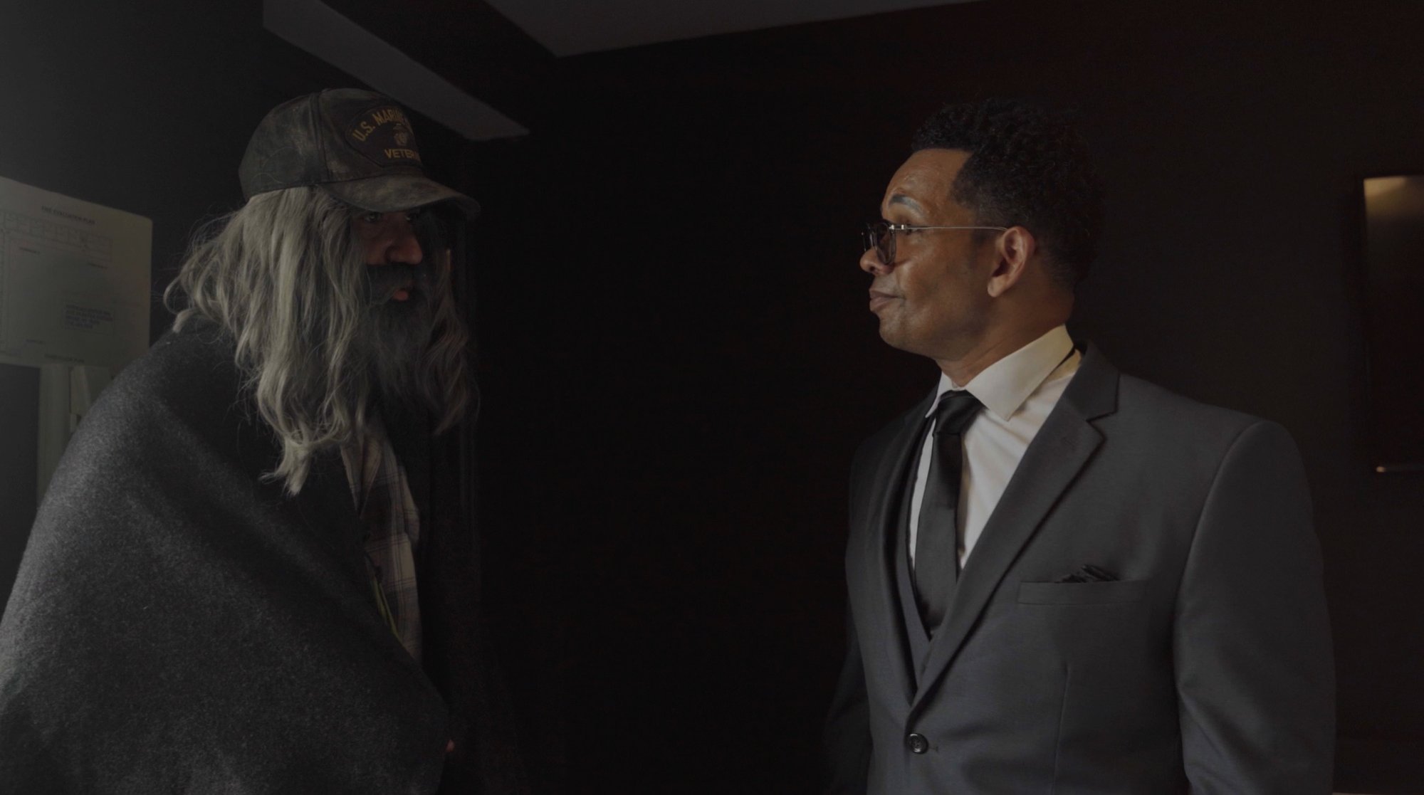 Still from The Veteran: Jose Torres, bearded and wrapped in a blanket under a U.S. Marines veteran cap, faces Maxwell Cade, a CEO in a grey suit, in a dark hallway.