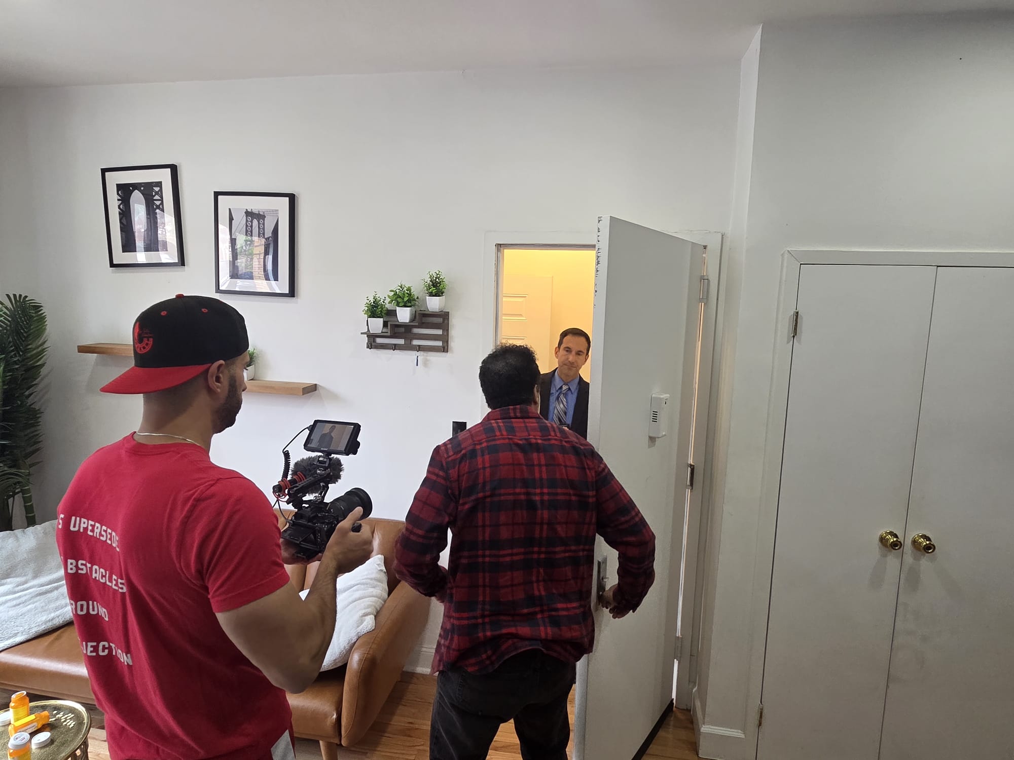 Wilfred La Salle, in a red T-shirt and black cap, films a scene for The Veteran in a Bushwick apartment as Yanko Valentin-Perez, in a red plaid shirt, opens a door to a man in a suit.