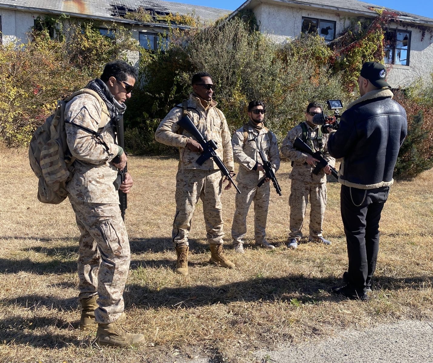 Wilfred La Salle, in a black cap and shearling jacket, films four actors in desert camouflage holding prop rifles in front of a boarded-up, vine-covered building.
