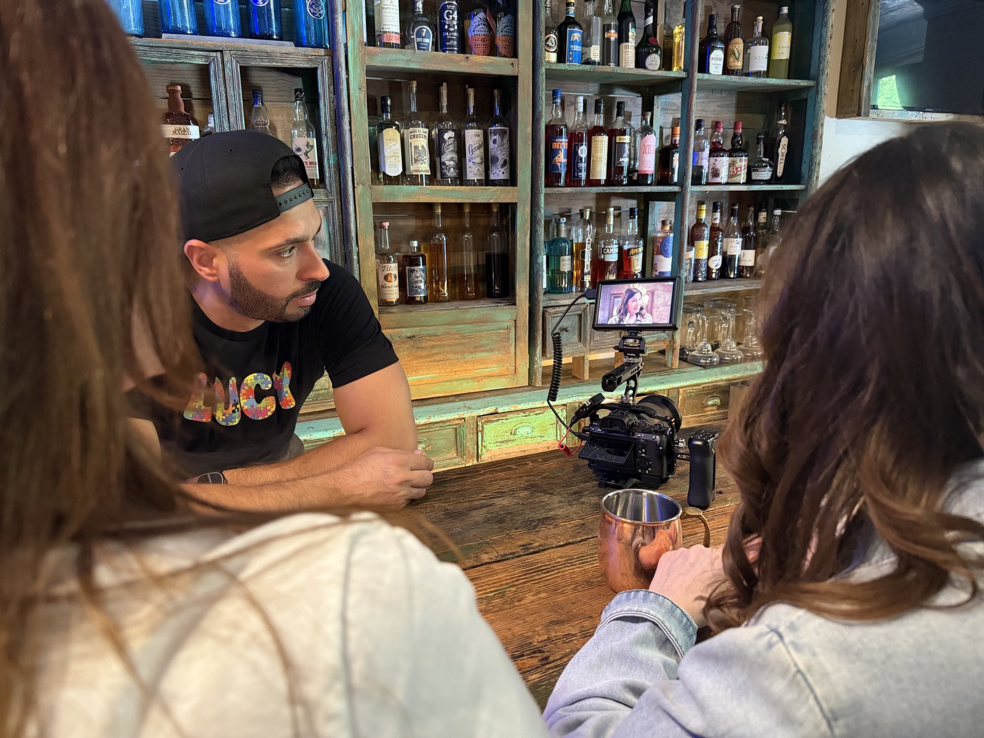 Wilfred La Salle, in a black LUCY T-shirt and backwards cap, leans on a bar lined with liquor bottles while a camera monitor shows the actress he is filming, as two women sit in the foreground.