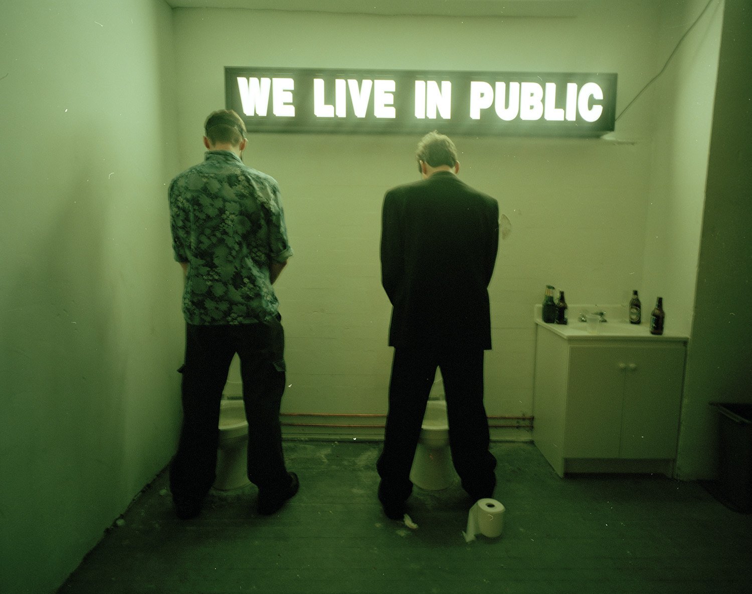 Two men stand before a neon sign reading We Live in Public in an archive photo
