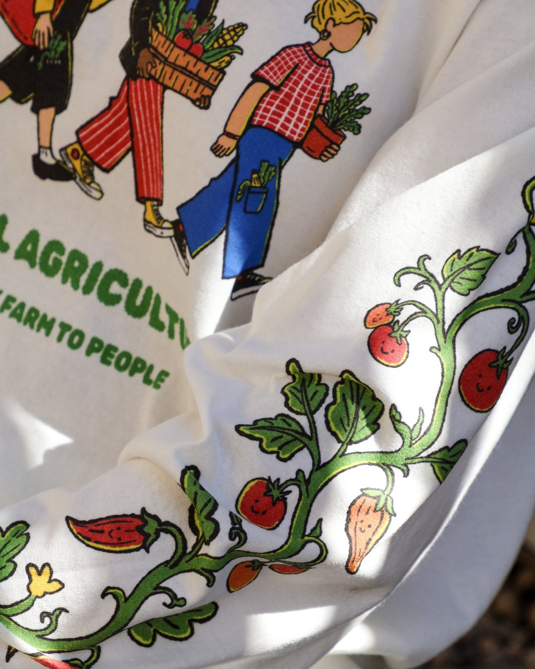 Close-up of the long-sleeve tee showing Fefi Castedo's line of people carrying produce and a tomato vine printed down the sleeve with the words Farm to People.