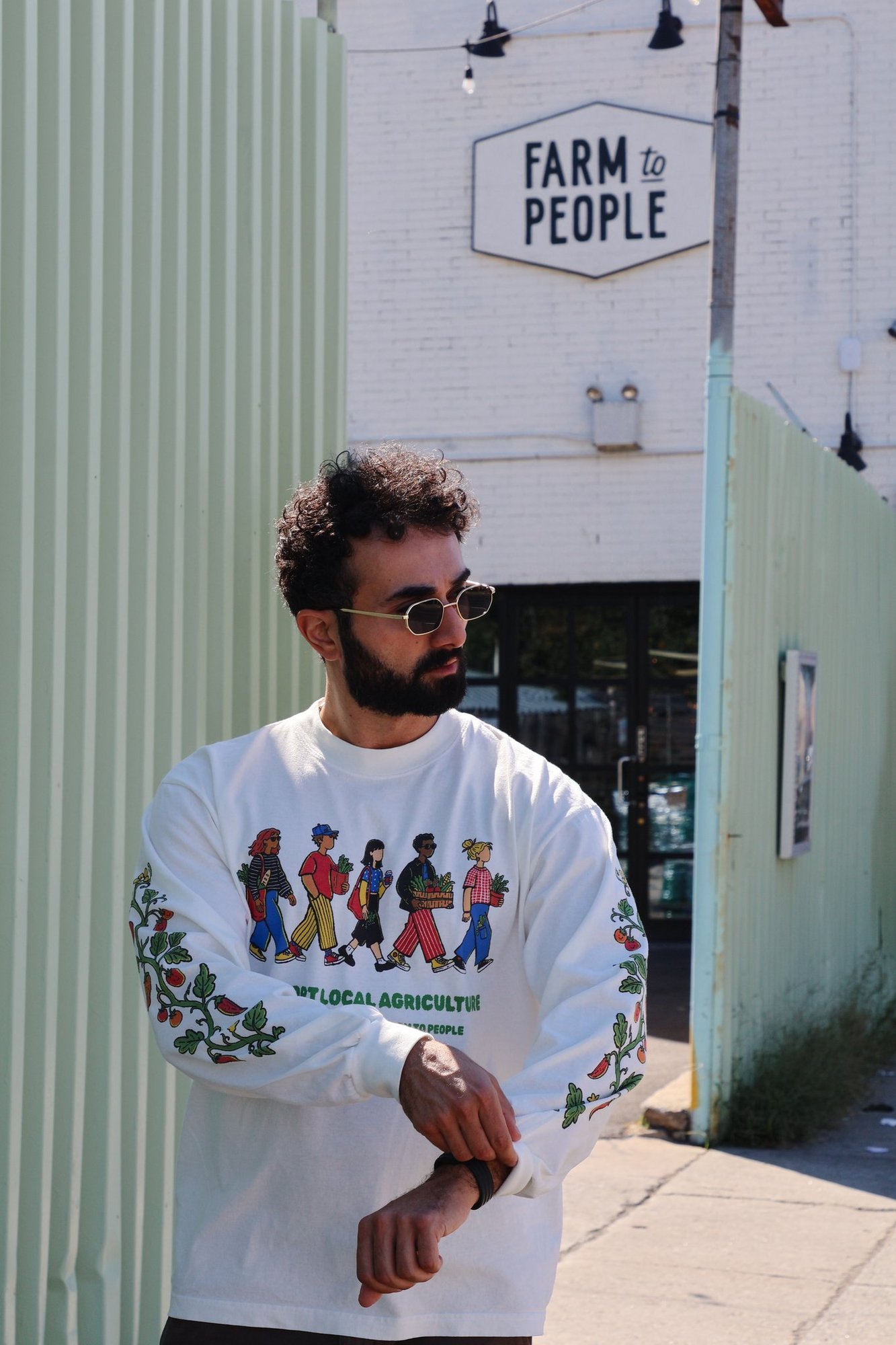 A man in the collection's long-sleeve tee stands on a sidewalk outside the Farm to People building, its sign visible above the entrance.