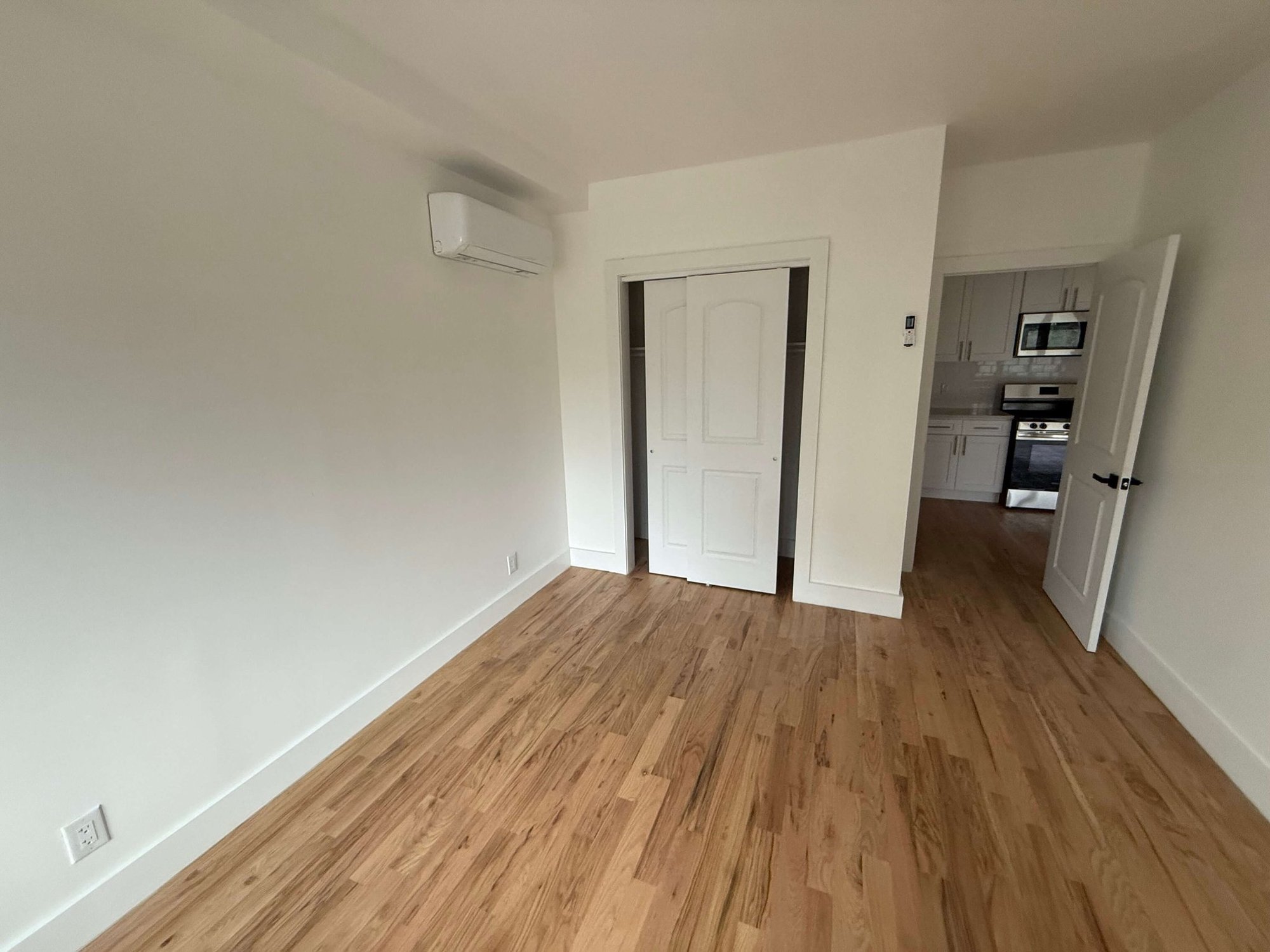 An empty bedroom with light hardwood floors, a wall-mounted split-system air conditioner, a closet with sliding doors and a view through to a white kitchen.