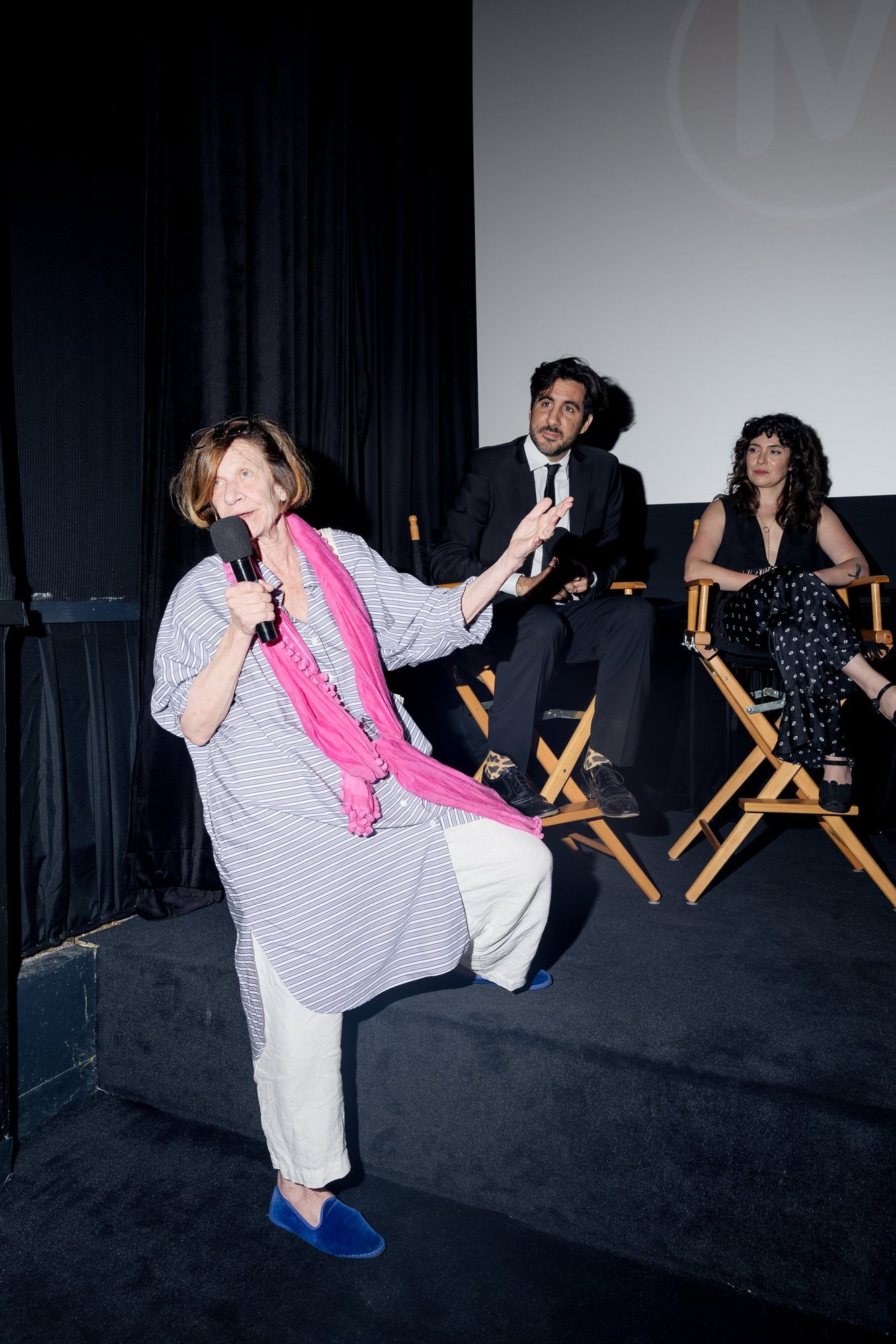 Brenda Cullerton, holding a microphone and wearing a pink scarf, speaks on a stage in front of a cinema screen while Anthony DiMieri and a woman sit in director's chairs behind her.